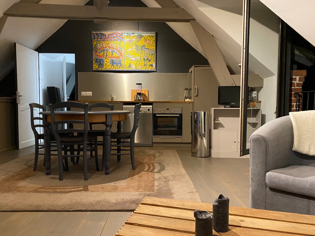 An open kitchen and dining area features a wooden table surrounded by six chairs. Modern appliances, including an oven and a refrigerator, are integrated within cabinetry. A colorful wall art piece adds a touch of character, while light from the windows accentuates the room's height and design.