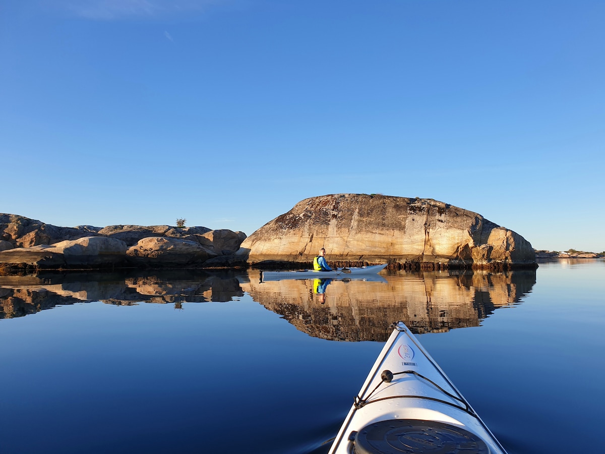 A calm, glassy water surface reflects a clear blue sky and the rugged outline of a large rock formation. A lone kayaker is seen paddling peacefully in the distance, surrounded by serene natural beauty.