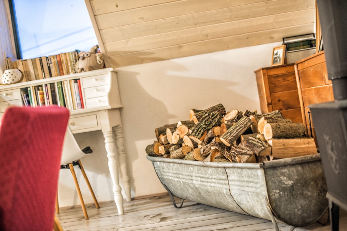 A rustic seating area features a unique metal tub filled with neatly stacked firewood, adding an element of warmth to the space. Natural light enters through large windows, illuminating a nearby desk with books and a wooden dresser.