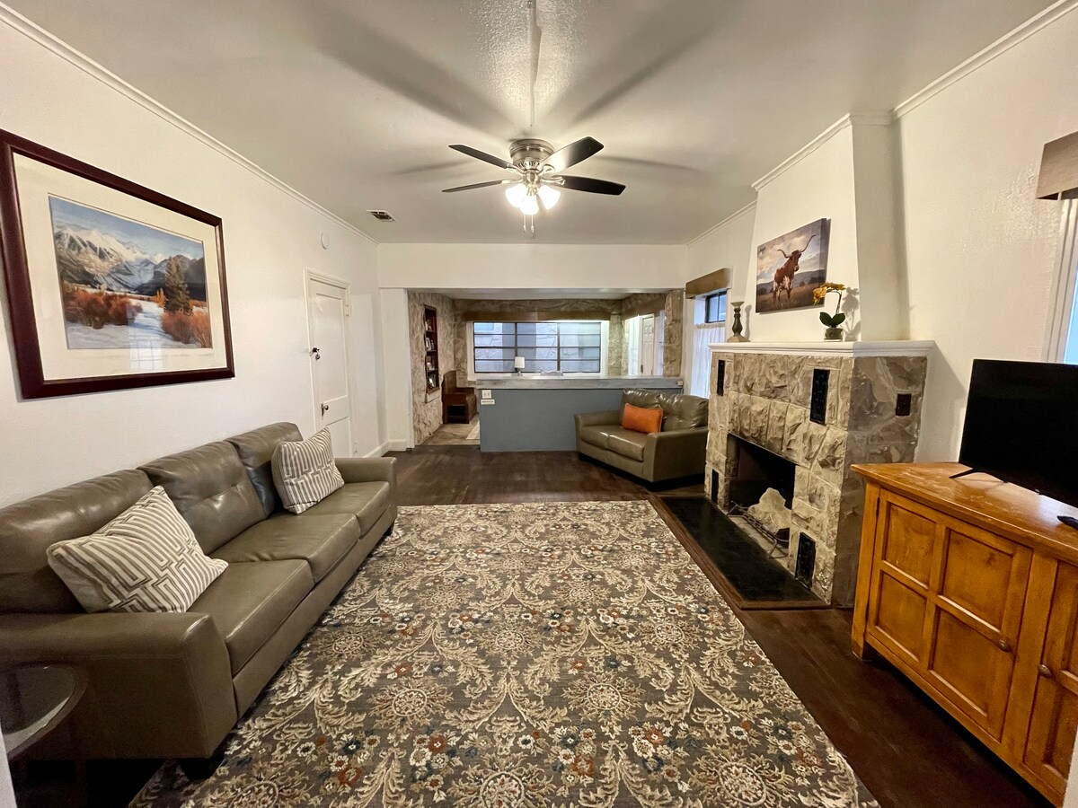 A spacious living room features a stone fireplace as its focal point. Two comfortable sofas are positioned around a large area rug, while natural light enters through multiple windows. A wooden cabinet stands against the wall, adding a functional element to the room.