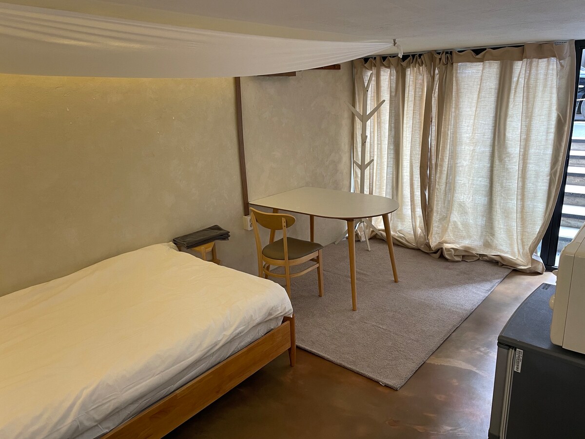 A minimalistic guest room features a spacious bed with white linens adjacent to a simple wooden table and chair. Natural light enters through soft curtains, illuminating the neutral-toned walls and floor. A cozy atmosphere is created with a clean and uncluttered design.
