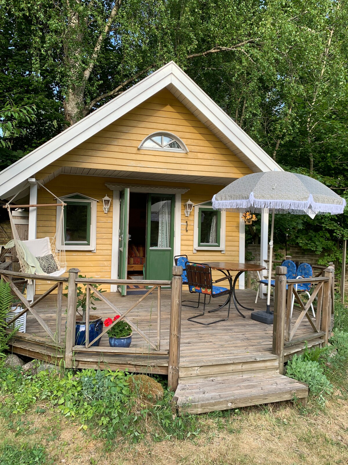 A charming cabin features a bright yellow exterior with white trim. A spacious wooden deck is adorned with a round table and chairs, accompanied by a decorative umbrella. Lush greenery surrounds the cabin, enhancing the inviting outdoor space.