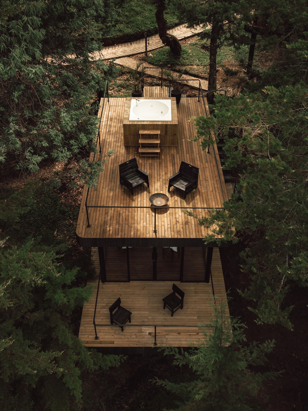 A modern cabin is nestled among trees, featuring a spacious wooden deck with four black chairs surrounding a circular fire pit. A hot tub is positioned on the deck, offering a secluded outdoor relaxation space. Paths winding through the forest can be seen nearby.