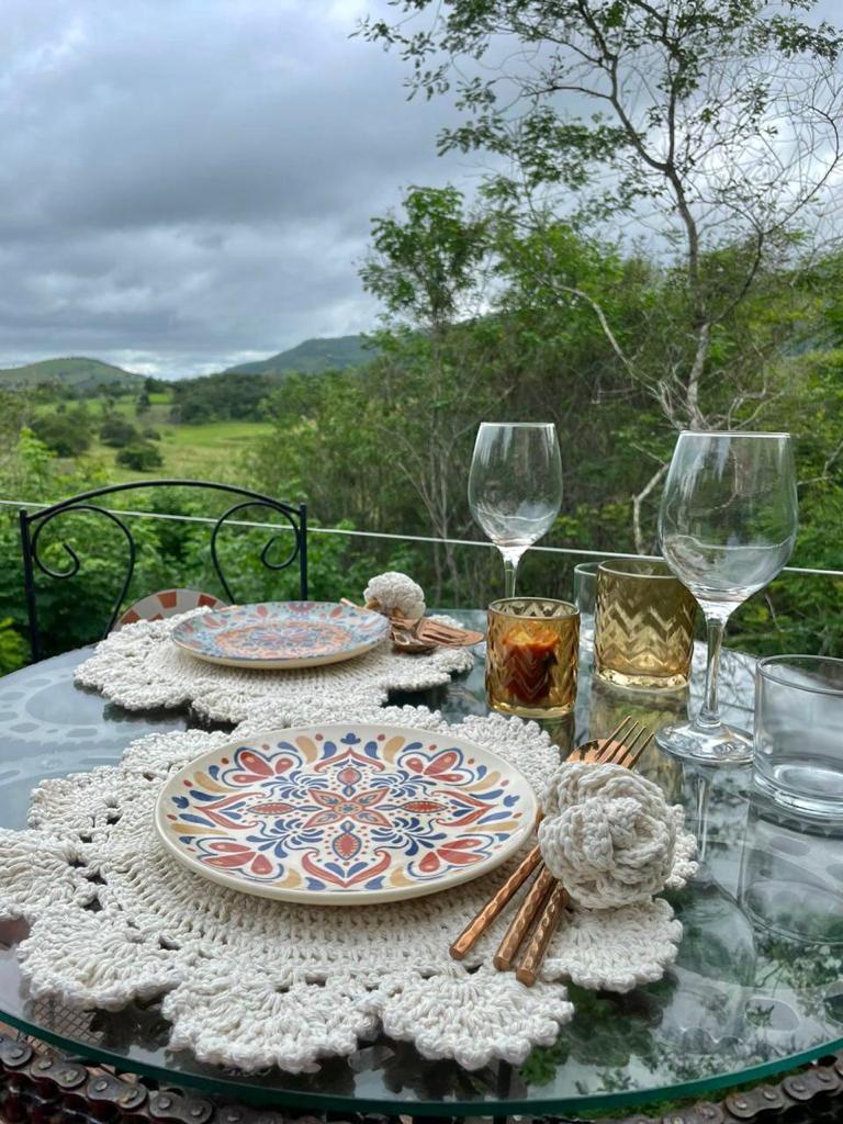 A dining table is arranged with patterned plates, elegant glassware, and decorative napkin holders. The setting features a handcrafted crochet tablecloth, with a backdrop of lush greenery and rolling hills under a cloudy sky, creating a serene and inviting outdoor dining atmosphere.