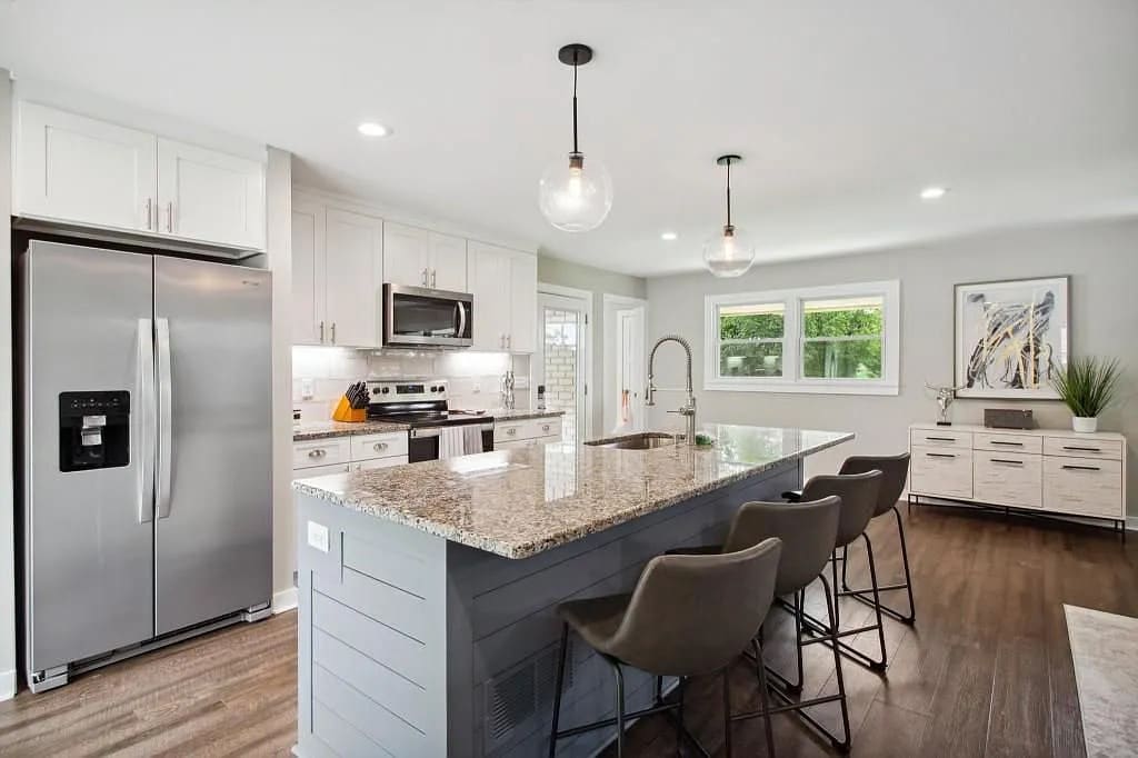 A modern kitchen features sleek stainless steel appliances, including a refrigerator and an oven, set against light-colored cabinetry. A spacious island adorned with granite countertops offers seating with four bar stools. Natural light floods in through large windows, illuminating the functional layout.