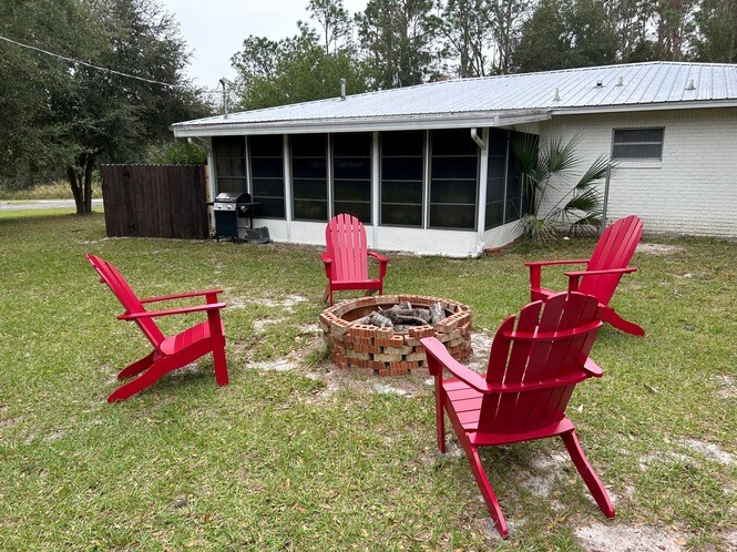 A cozy outdoor fire pit area is surrounded by four bright red Adirondack chairs arranged in a circular formation. The fire pit features a brick border and is set on a grassy lawn, providing a comfortable space for gatherings. A screened-in porch is visible in the background.
