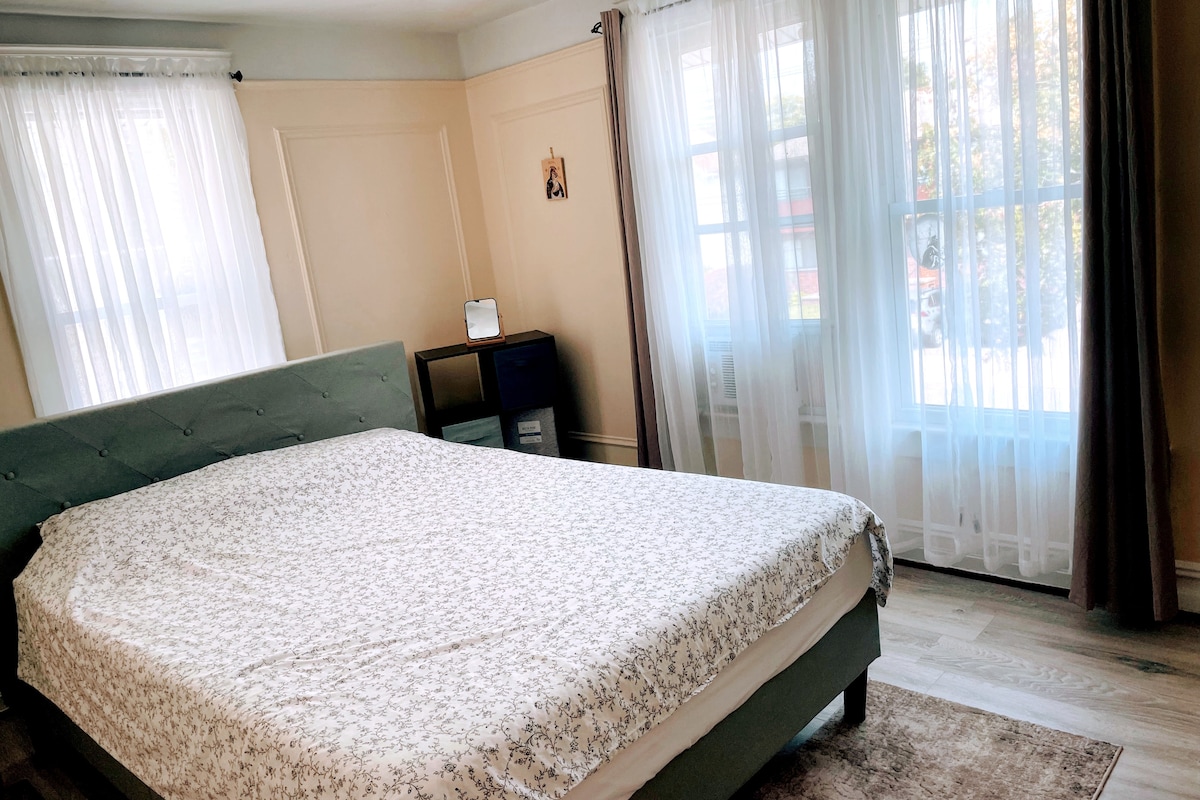 A spacious guest bedroom features a large green bed with a light-colored patterned coverlet. Two windows allow natural light to fill the room, framed by sheer white curtains, while a small nightstand is placed beside the bed against a softly colored wall.