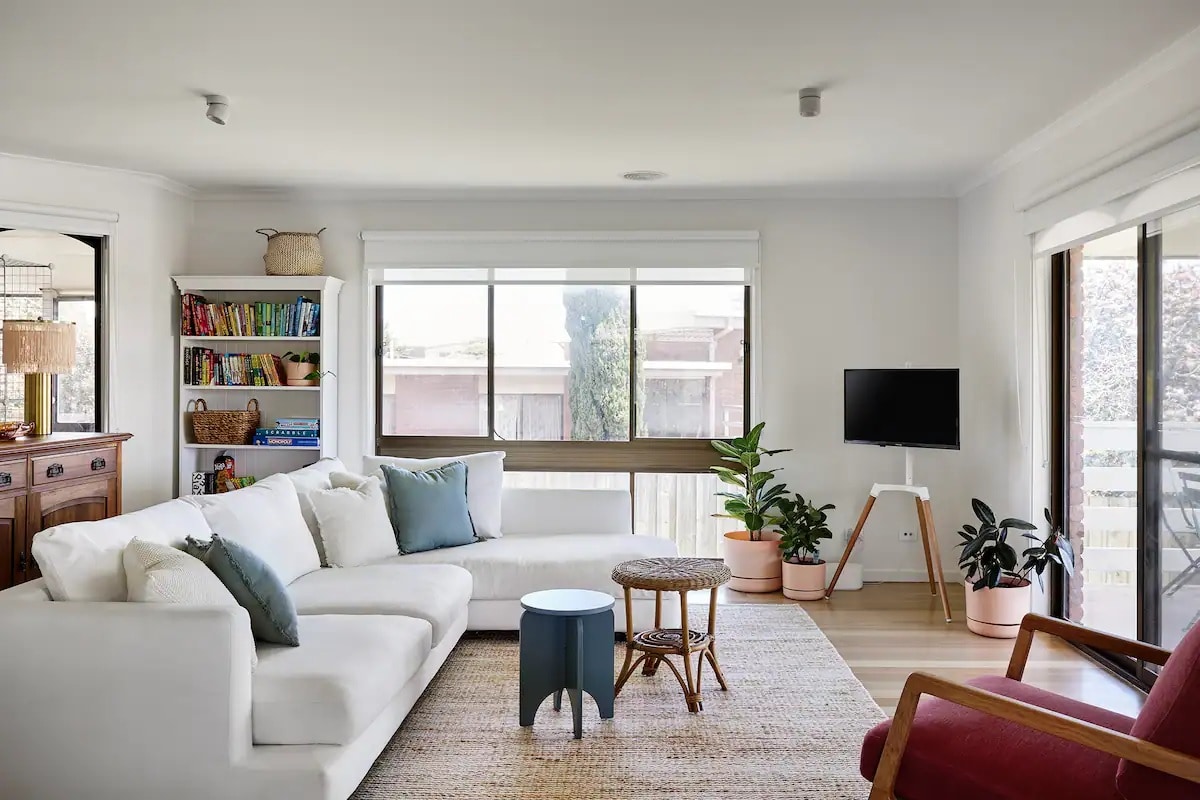 A bright and airy living area features a spacious white sofa adorned with teal accent pillows, complemented by a wooden side table. Large windows allow natural light to illuminate the room, while indoor plants add a touch of greenery. A small TV is mounted on a wall opposite a reading nook.
