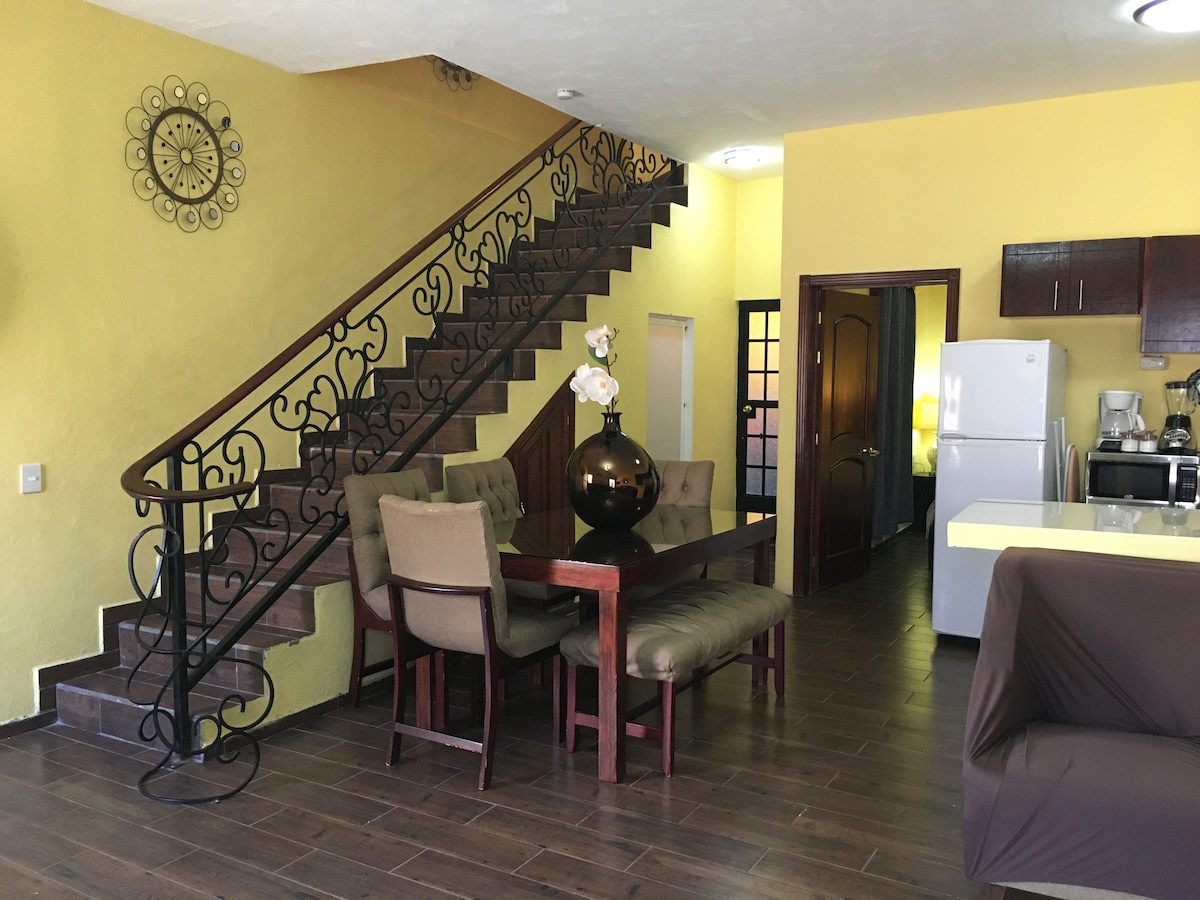 A dining area is showcased with a wooden table surrounded by upholstered chairs. A decorative vase with white flowers sits at the center. A stylish staircase with an ornate iron railing leads to the upper level, while a glimpse of the kitchen and refrigerator is visible in the background.