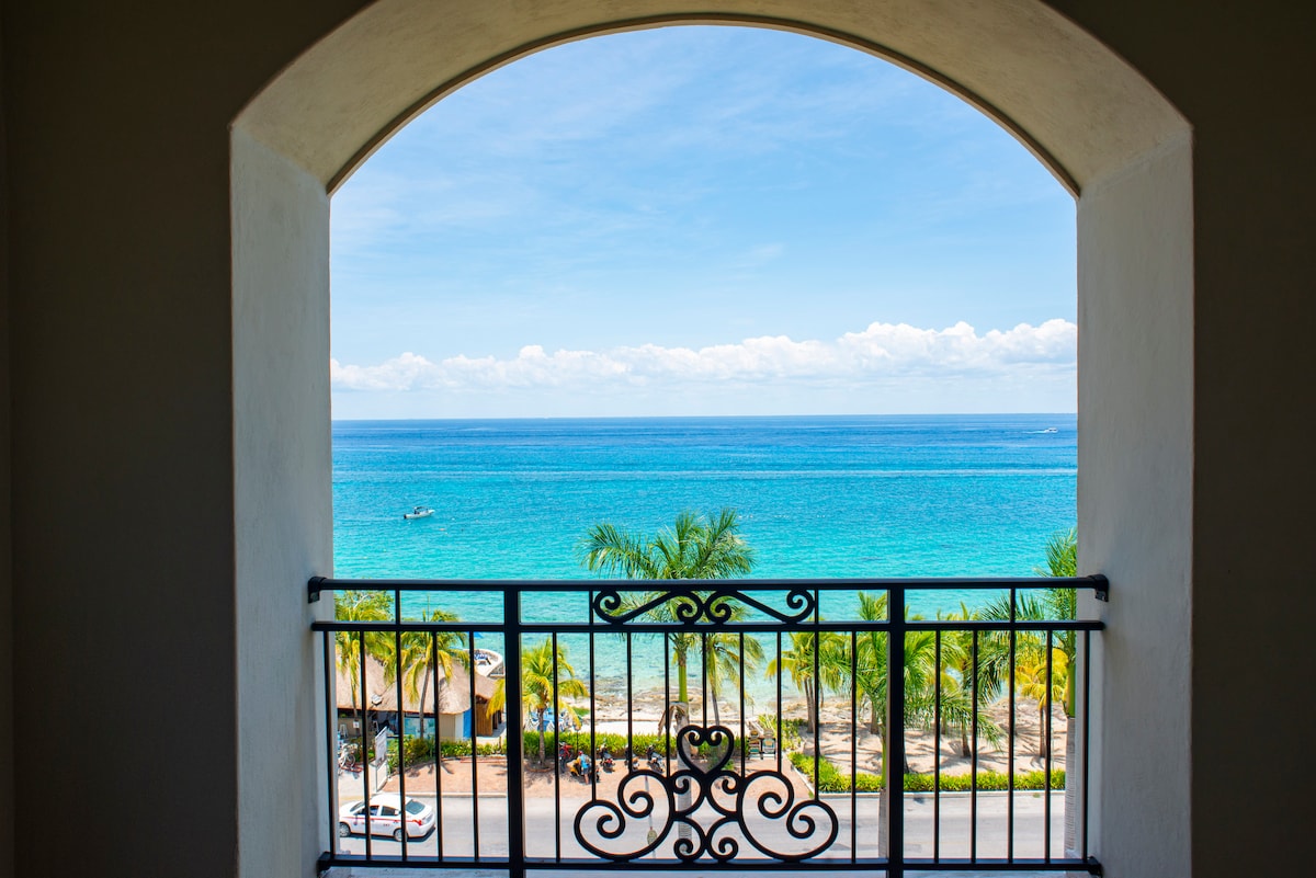 An ocean view is framed by a stylish wrought iron balcony railing, revealing vibrant turquoise waters and palm trees swaying gently in the breeze. A clear blue sky with scattered clouds enhances the serene coastal setting.