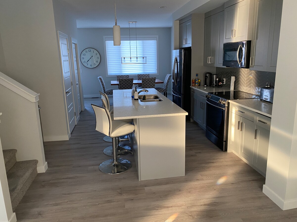 A modern kitchen features quartz countertops and stainless steel appliances, including a refrigerator and stove. Bright lighting highlights a central island with seating for four. A dining area is visible in the background, complemented by a large wall clock and stylish chairs.