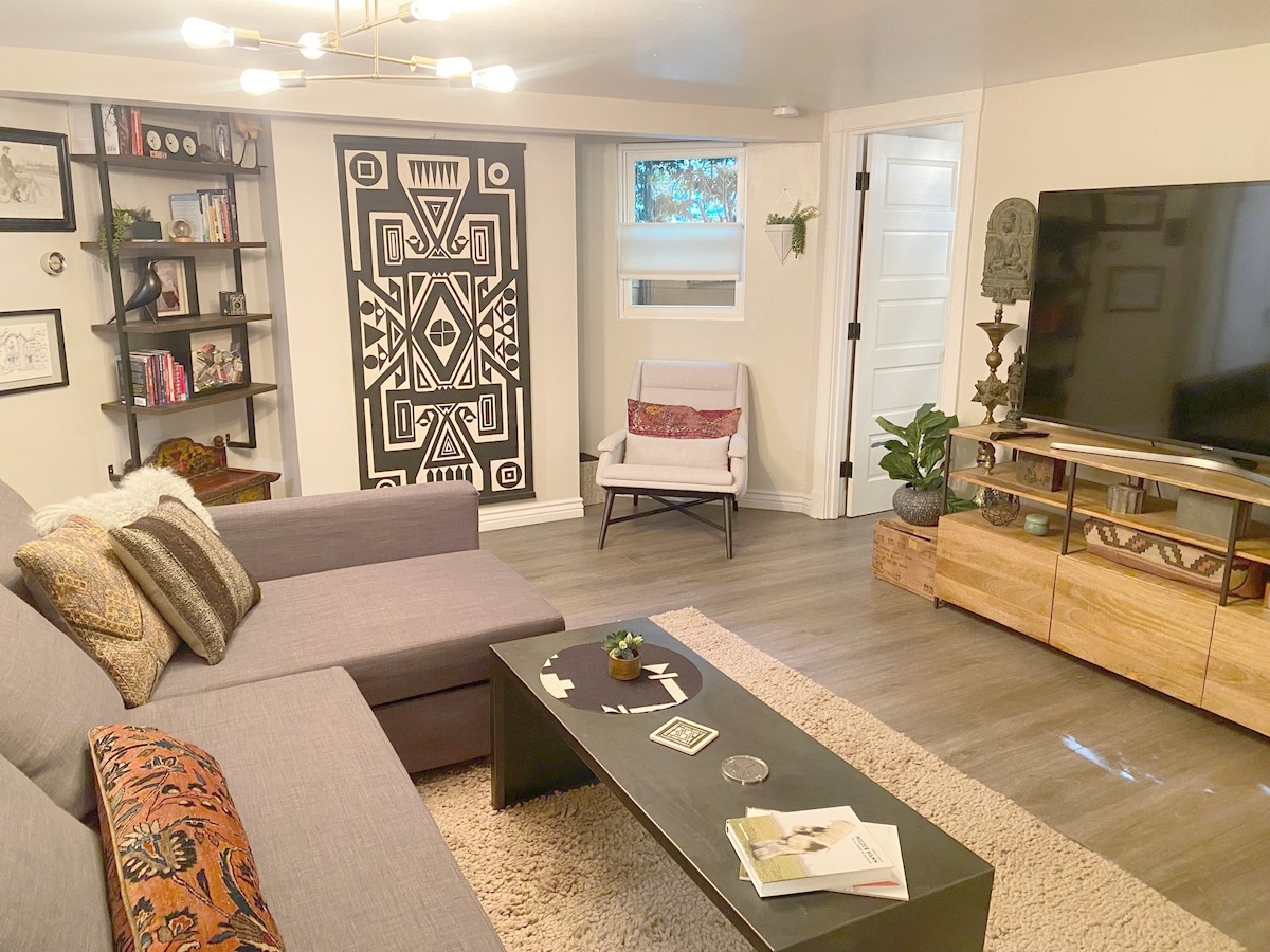 The living room features a sectional sofa in gray upholstery, complemented by a colorful accent pillow. A coffee table sits centrally with decor items. A wall-mounted TV is positioned opposite the sofa, next to a decorative shelf filled with books and art. A large area rug adds warmth.