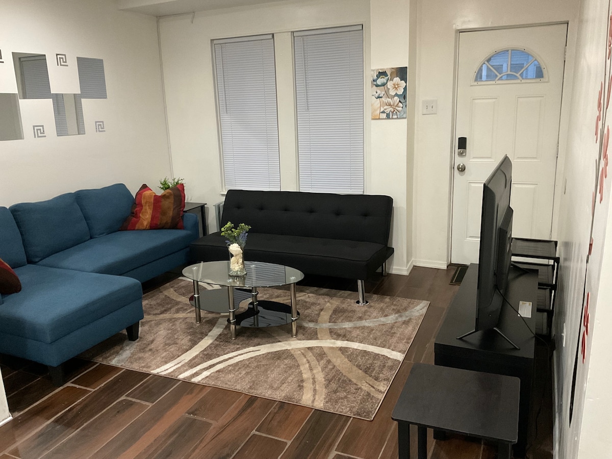 A cozy living area features a blue sectional couch and a black futon, arranged around a glass coffee table on a patterned area rug. A flat-screen television sits on a black stand, with natural light filtering through two window treatments.