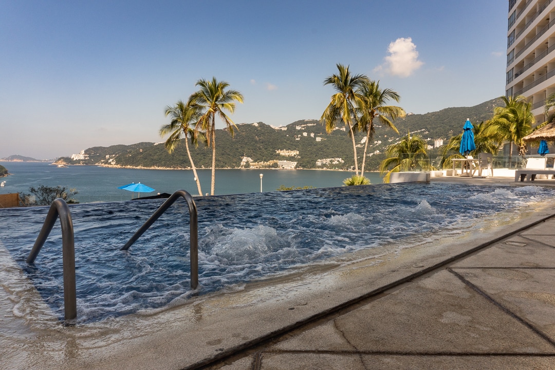 An infinity pool overlooks a scenic bay, framed by palm trees swaying gently in the breeze. Clear blue water reflects the sky, while the surrounding paved area includes lounge chairs and umbrellas, ensuring a relaxing environment for guests.