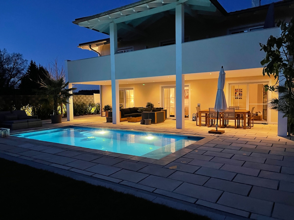 The tranquil outdoor area is illuminated at night, featuring a private pool surrounded by comfortable seating and a dining table. The spacious terrace is adorned with potted plants, creating a serene ambiance in a well-maintained garden.