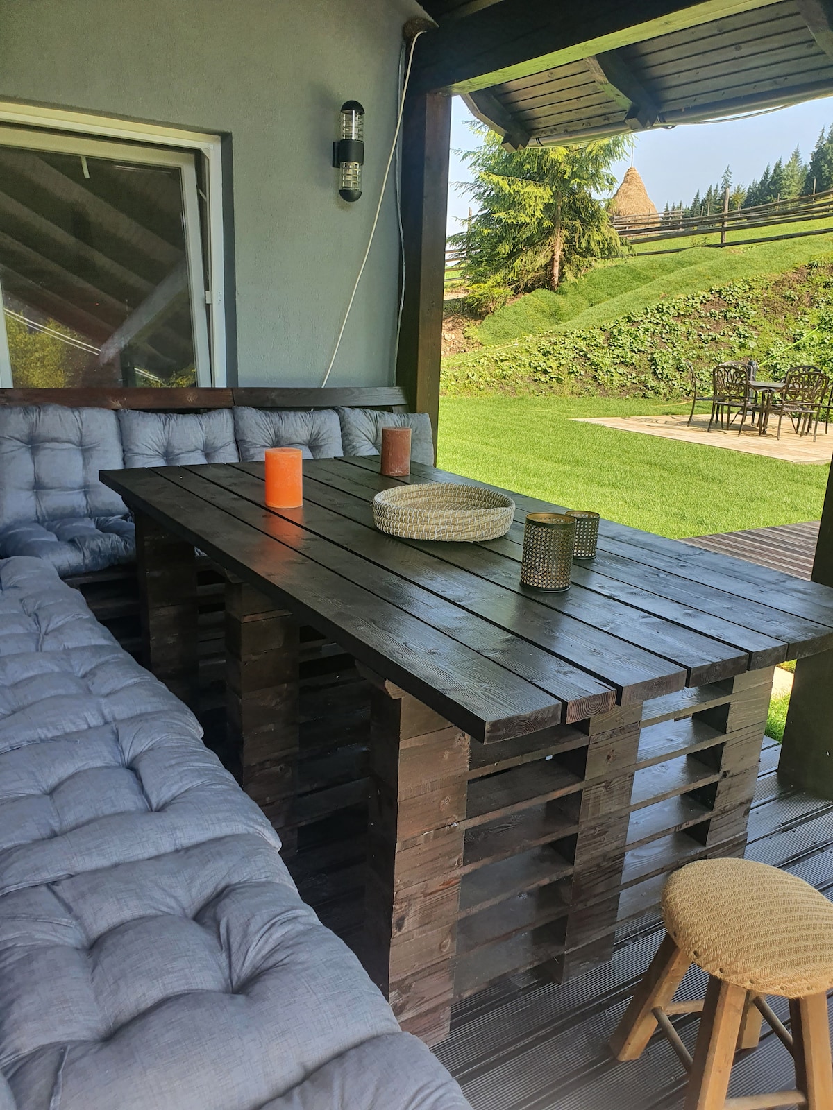 An outdoor seating area is presented with a wooden table and cushions. Various decorative items, including candles and woven baskets, are placed on the table. Lush green grass and forested hills are visible in the background, creating a serene outdoor environment.