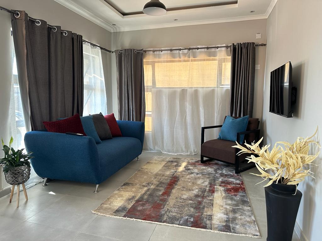 A cozy lounge area features a blue sofa paired with accent pillows in deep red. A dark wood chair complements the modern decor. Natural light filters through sheer curtains, illuminating a textured area rug and a decorative plant in a pot.