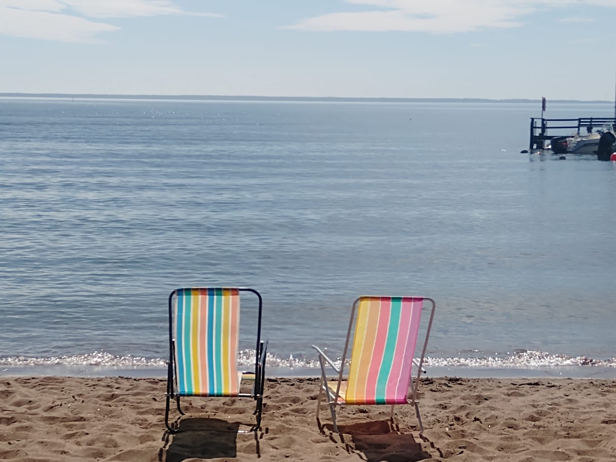 Two colorful beach chairs with striped patterns are positioned on a sandy beach, facing the calm water of the lake. The gentle waves lap at the shore, while a dock is visible in the distance under a clear blue sky.