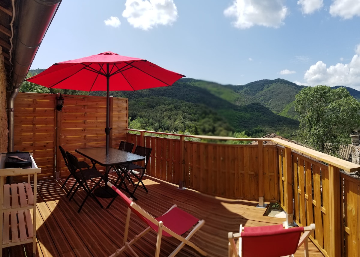 A spacious wooden terrace features a large red umbrella providing shade over a dining table and chairs. The surrounding mountains are visible in the background under a clear blue sky, enhancing the outdoor setting. A couple of lounge chairs are positioned for relaxation.