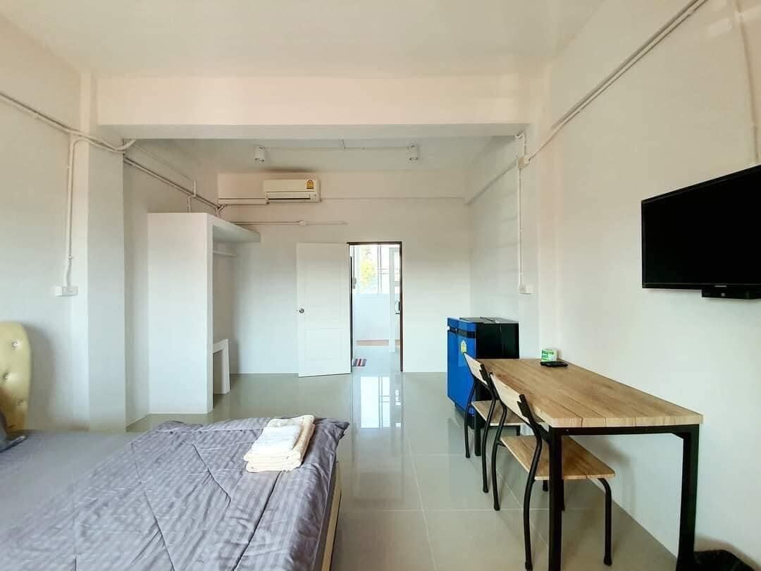 The room features a spacious bed with light bedding, complemented by a wooden table and bench set. A wall-mounted television is positioned opposite the bed, and a small kitchenette area with a blue cabinet is visible. Natural light enters through the door leading to the balcony.