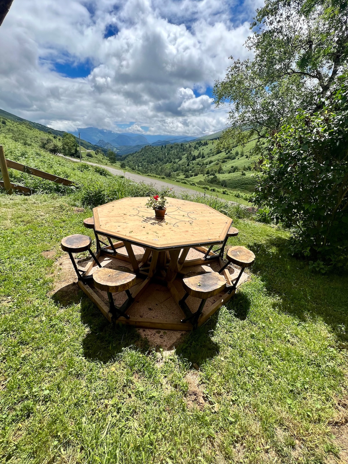 A rustic outdoor table with a round top made of wood is surrounded by six wooden stools. The setting offers a view of rolling hills and a winding road in the distance, under a partly cloudy sky. Green grass surrounds the table, adding to the serene atmosphere.