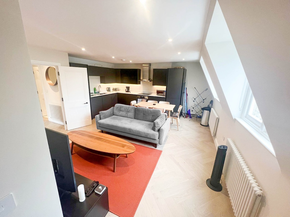 A comfortable living space features a gray sofa and a wooden coffee table atop a red area rug. A dining table is set in the background, with a fully equipped kitchen nearby. Light filters through the window, highlighting the apartment's modern design.