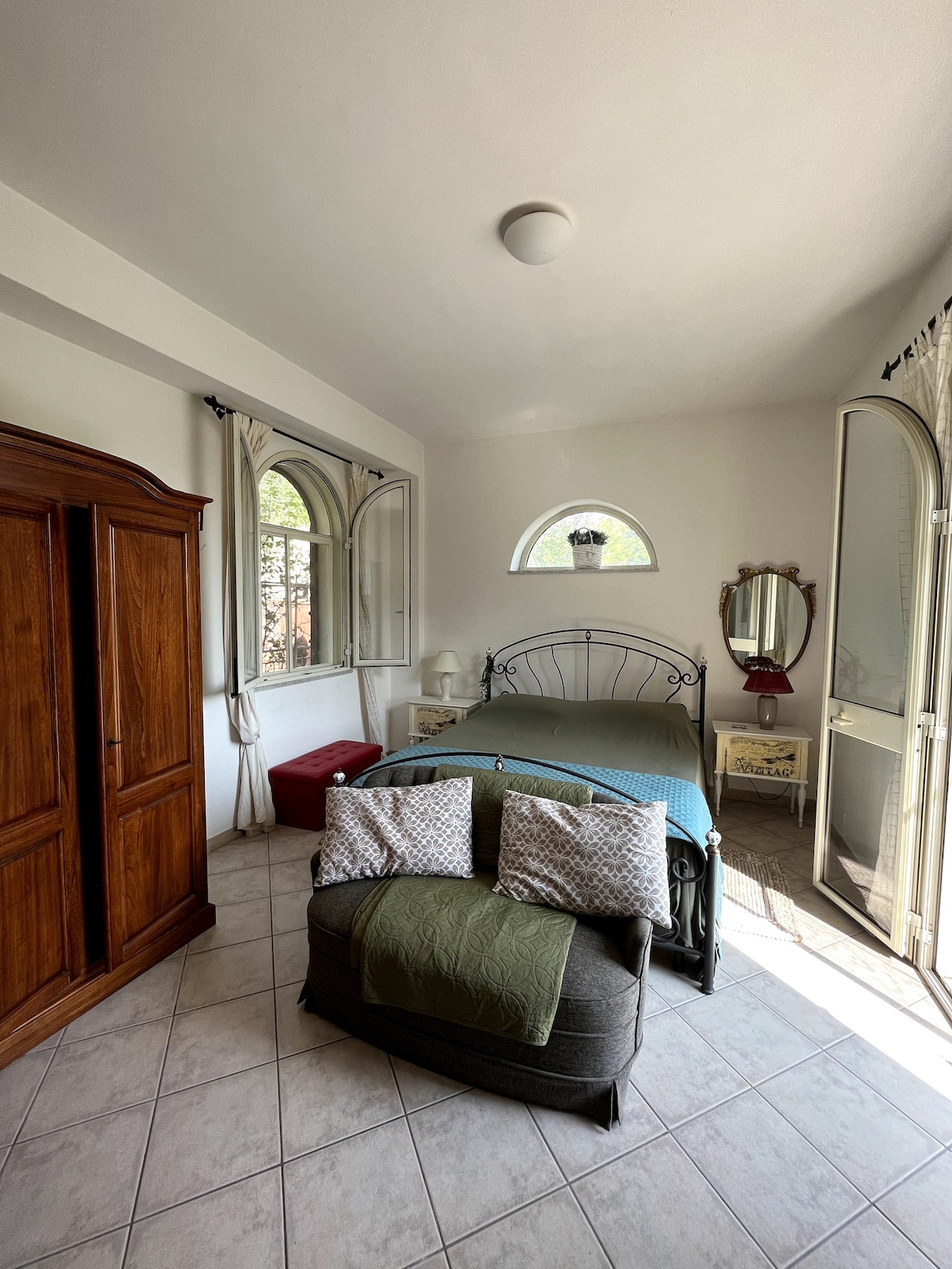 A bright bedroom features a spacious bed with soft bedding and two large windows allowing natural light. A wooden wardrobe stands against the wall, complemented by a decorative mirror. A cozy seating area is created with two plush chairs positioned near a window.