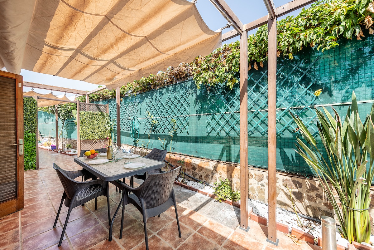 An outdoor dining area features a table surrounded by four chairs, set beneath a sunshade. Lush greenery borders the space, providing a sense of privacy. The tile flooring complements the serene environment, ideal for enjoying meals al fresco.