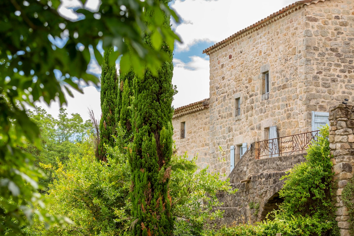The stone façade of the traditional building is framed by lush greenery, including tall cypress trees. A balcony with decorative railing extends from the upper level, providing an inviting view of the surrounding nature.