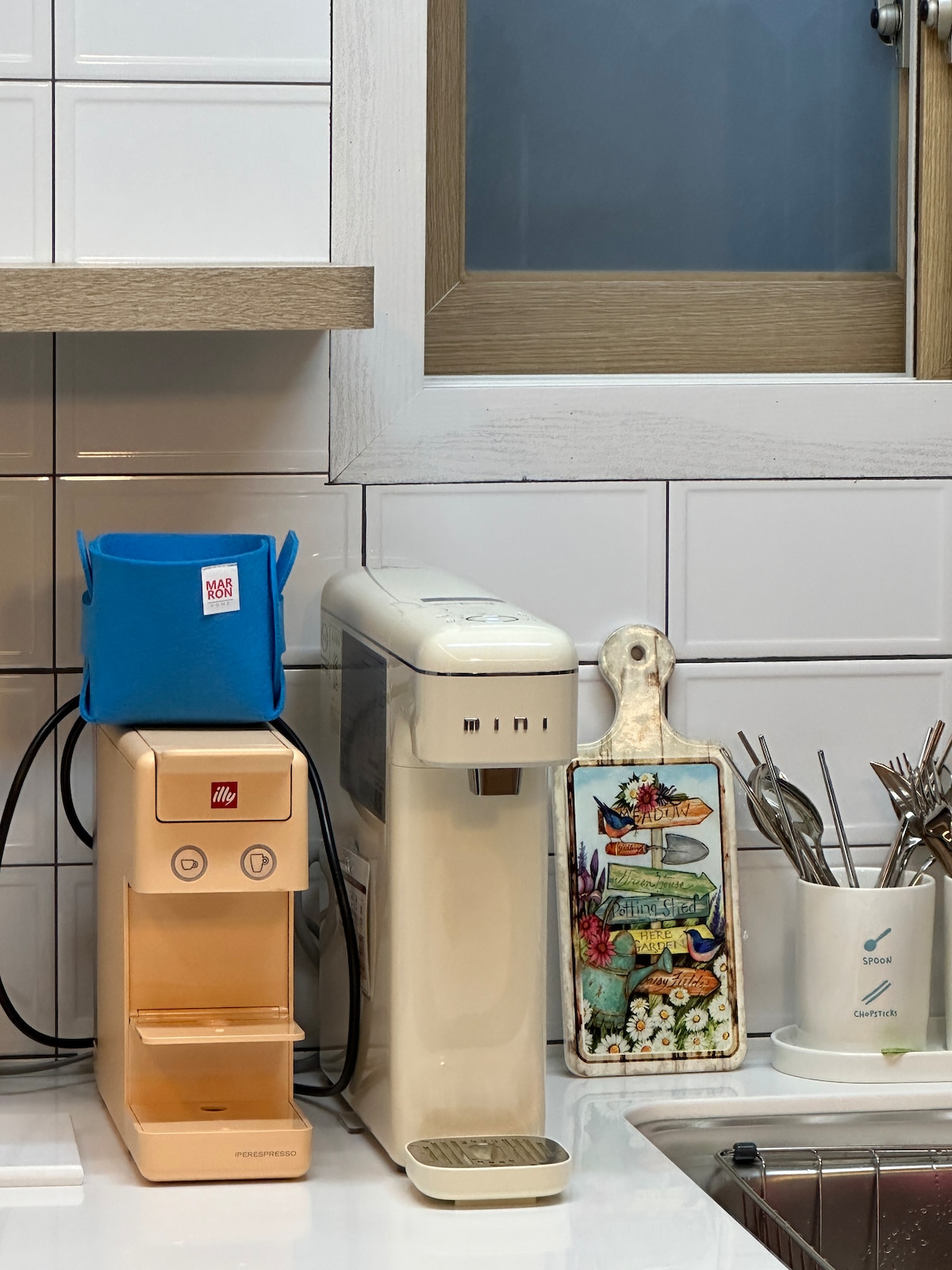 A kitchen countertop features a coffee machine in a soft peach color and a sleek water purifier in white. A decorative cutting board displaying colorful sea-themed art is positioned next to them, alongside a holder filled with utensils.