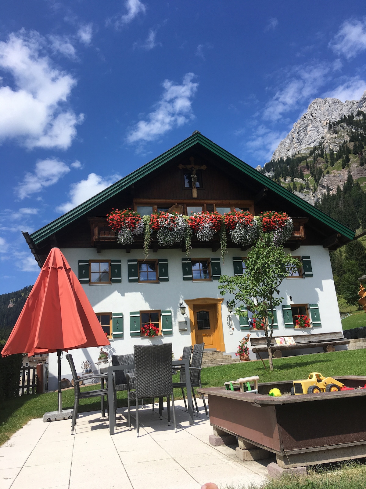A traditional alpine farmhouse displays vibrant flower boxes under a clear blue sky. A patio area is equipped with a table and chairs, complemented by a red umbrella. A children's play area is visible, along with scenic mountain views in the background.