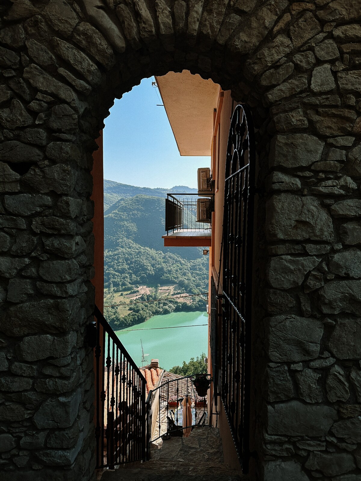 An arched stone doorway frames a scenic view of the turquoise Lago del Turano, surrounded by lush green mountains. A wrought iron gate completes the entrance, enhancing the tranquil atmosphere of the surrounding area.