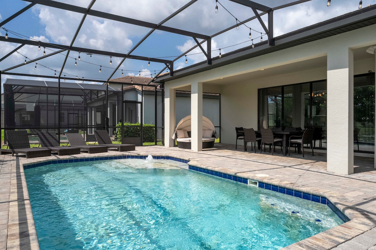 An inviting outdoor space features a large private pool surrounded by a spacious patio. Comfortable seating options are provided, along with a shaded area for relaxation. String lights enhance the ambiance, and the pool is enclosed by a screened-in structure, offering a sense of privacy.