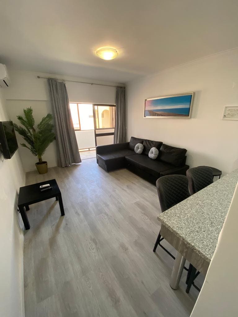 A living area is presented, featuring a dark sofa with two decorative pillows. A small table sits beside it, while a compact dining area with two charis is visible in the foreground. Natural light enters through a sliding glass door, revealing a view of an outdoor space.