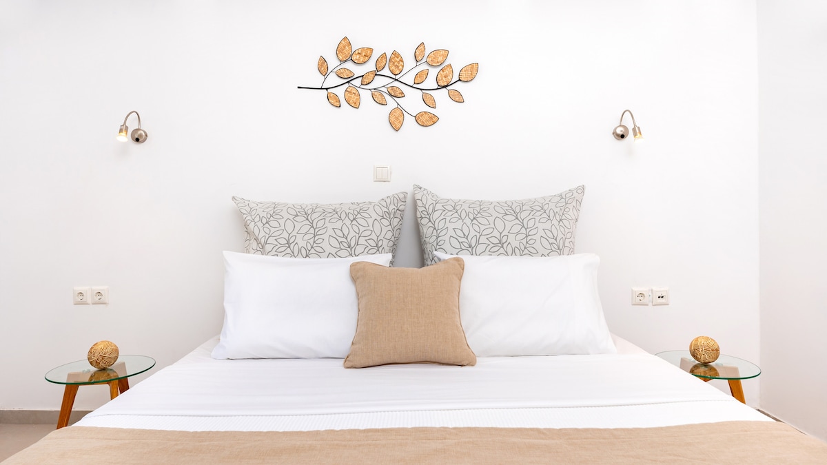 A well-appointed bedroom features a spacious bed with decorative pillows and a neutral-toned accent cushion. Two round bedside tables with decorative stones are positioned on either side, while a leaf-patterned wall art piece adds a subtle touch of elegance above the bed.