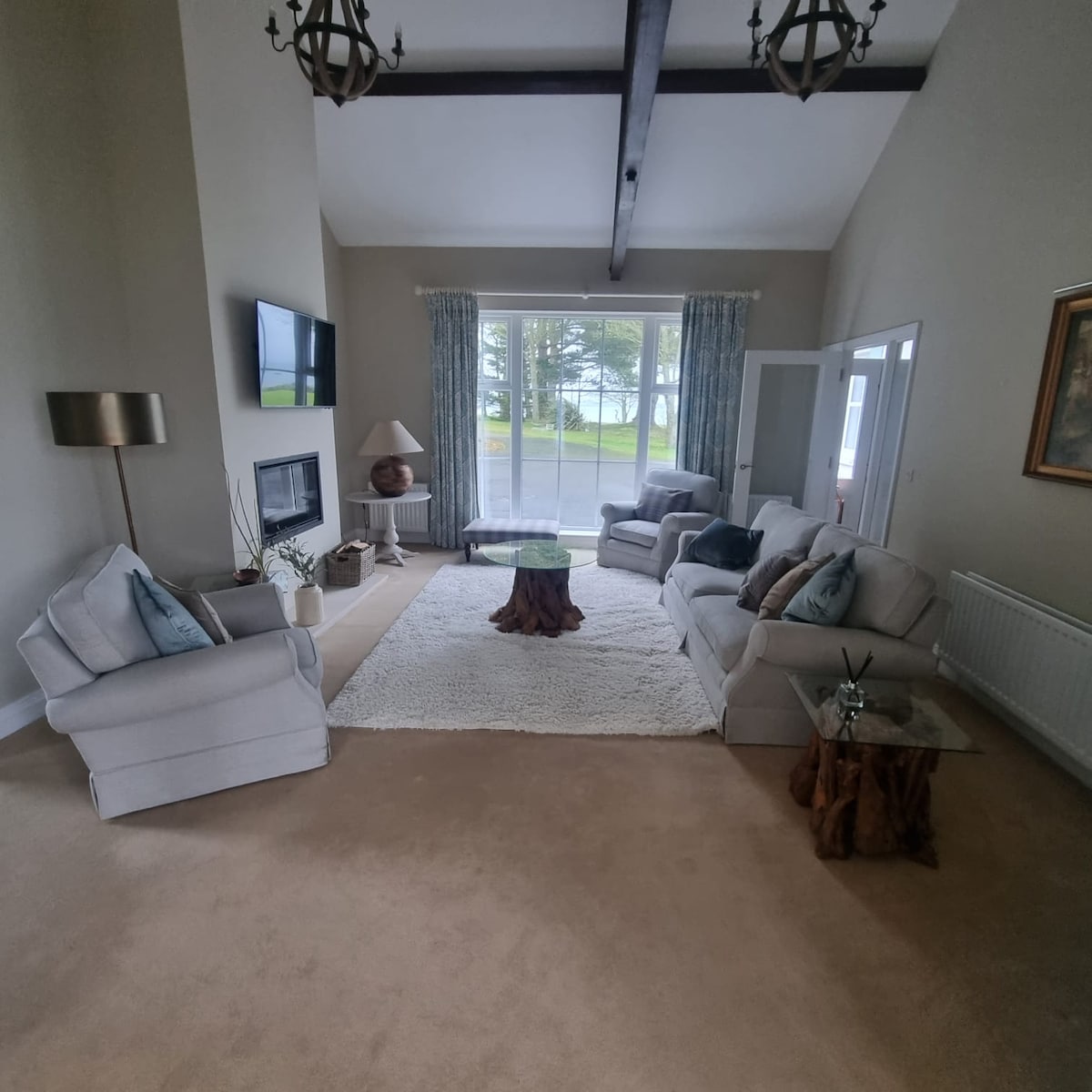 A spacious living area features large windows providing views of the outdoors. Comfortable seating arrangements are highlighted by a central area rug. Natural light fills the room, accentuating the soft color palette and decorative elements like a coffee table and floor lamp.