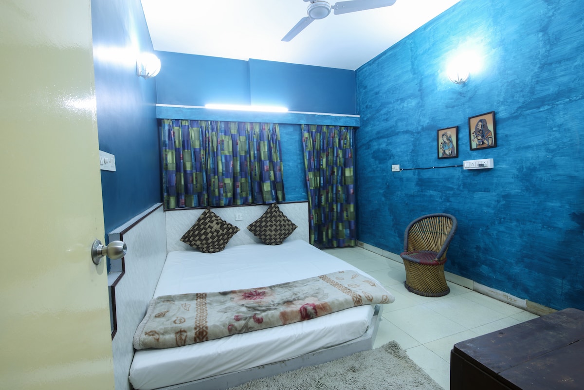 A cozy bedroom features a spacious bed with a soft blanket and patterned throw. The walls are painted in a vibrant blue hue, complemented by colorful curtains. A wicker chair is placed beside a wooden table, and framed artwork adorns the wall.