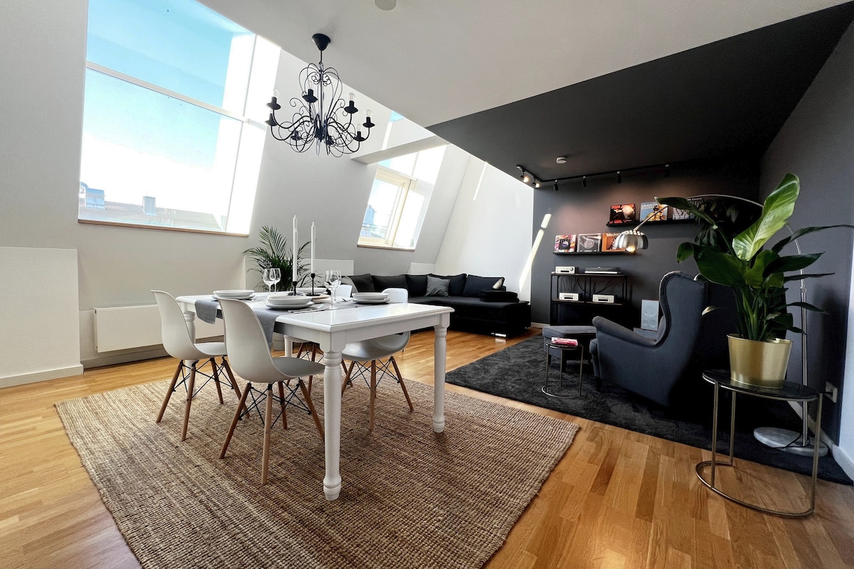 The stylish living area features a dining table surrounded by six white chairs, complemented by a cozy seating area with dark upholstery. A large window allows natural light to illuminate the space. Decorative plants and a modern chandelier add to the contemporary decor.