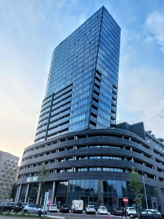 A modern high-rise building features a sleek glass façade and distinct angular design. The multi-level structure is complemented by a curved base and landscaped areas with small trees, providing an inviting entrance. Vehicles are parked along the street in the foreground.