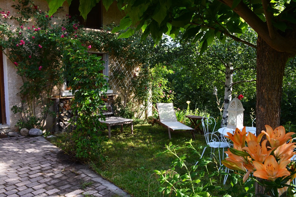 A serene outdoor space features a well-kept garden with lush greenery and vibrant flowers. Comfortable lounge chairs are positioned on a grassy area, accompanied by a small seating arrangement with white chairs. A stone pathway leads to the entrance of the house.