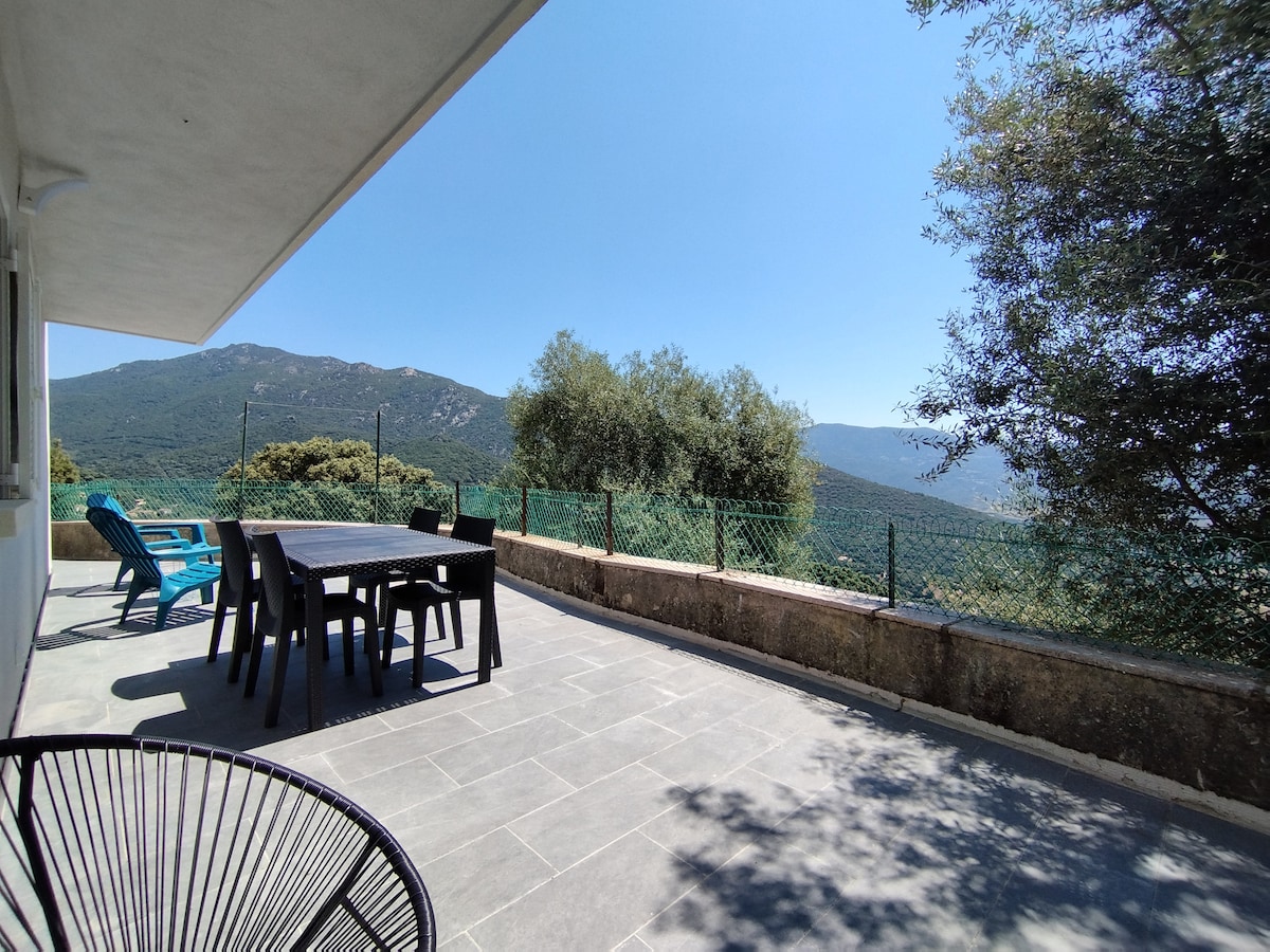 An outdoor terrace features a large dining table surrounded by chairs, overlooking a scenic view of green hills and distant mountains. The space is complemented by a few lounge chairs, offering comfortable seating for relaxation. A surrounding fence provides a sense of privacy.