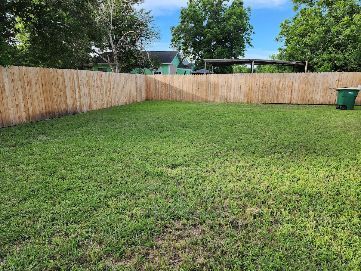 A spacious fenced backyard features a lush green lawn, offering ample room for outdoor activities. The area is bordered by wooden fencing, providing privacy and a safe environment. A green trash bin is visible in one corner of the yard.