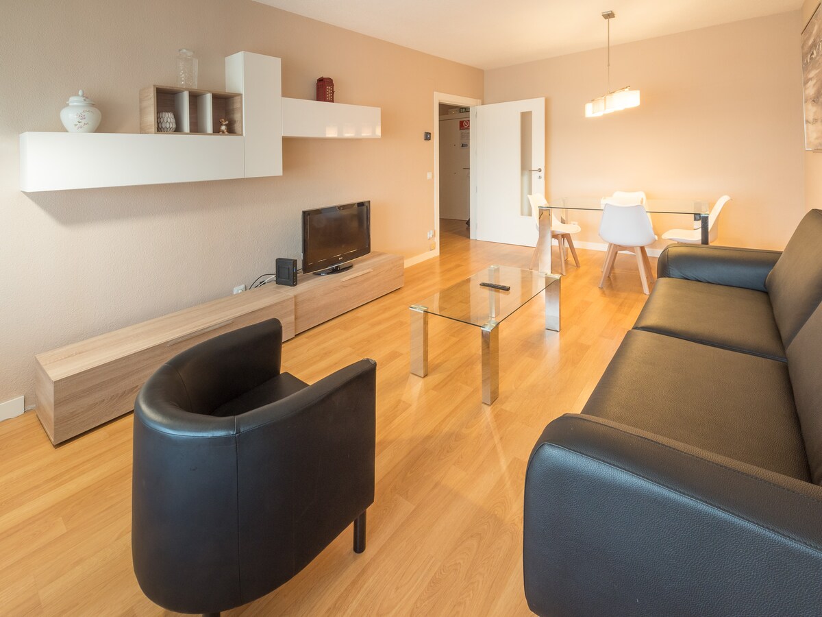 A modern living area features a sleek black sofa and two matching armchairs. A glass coffee table sits in the center, while a low wooden unit holds decorative items. A dining table with white chairs is visible in the background, complemented by soft lighting overhead.
