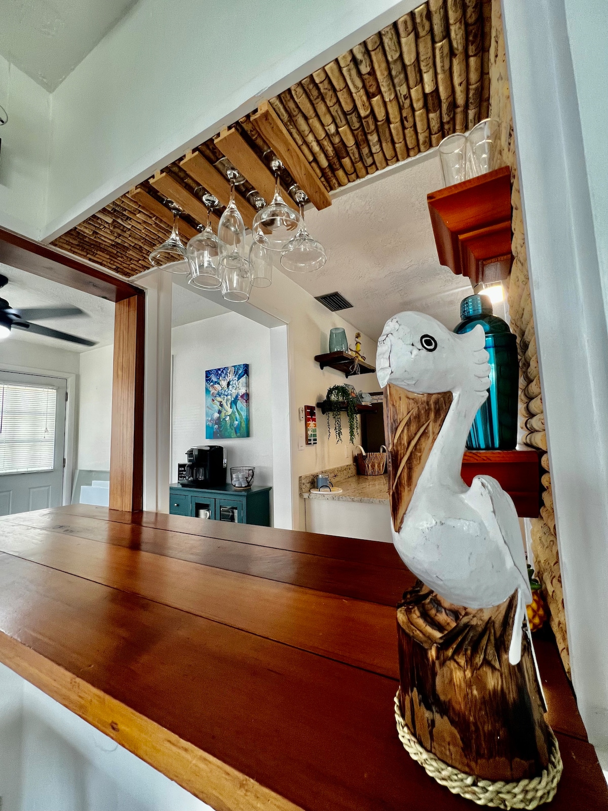 A handcrafted wooden pelican figurine stands prominently on the countertop, adding a coastal touch. Above, pendant lights hang beneath a bamboo ceiling, providing a warm glow. The background reveals glimpses of a colorfully decorated space with various shelves and artwork.