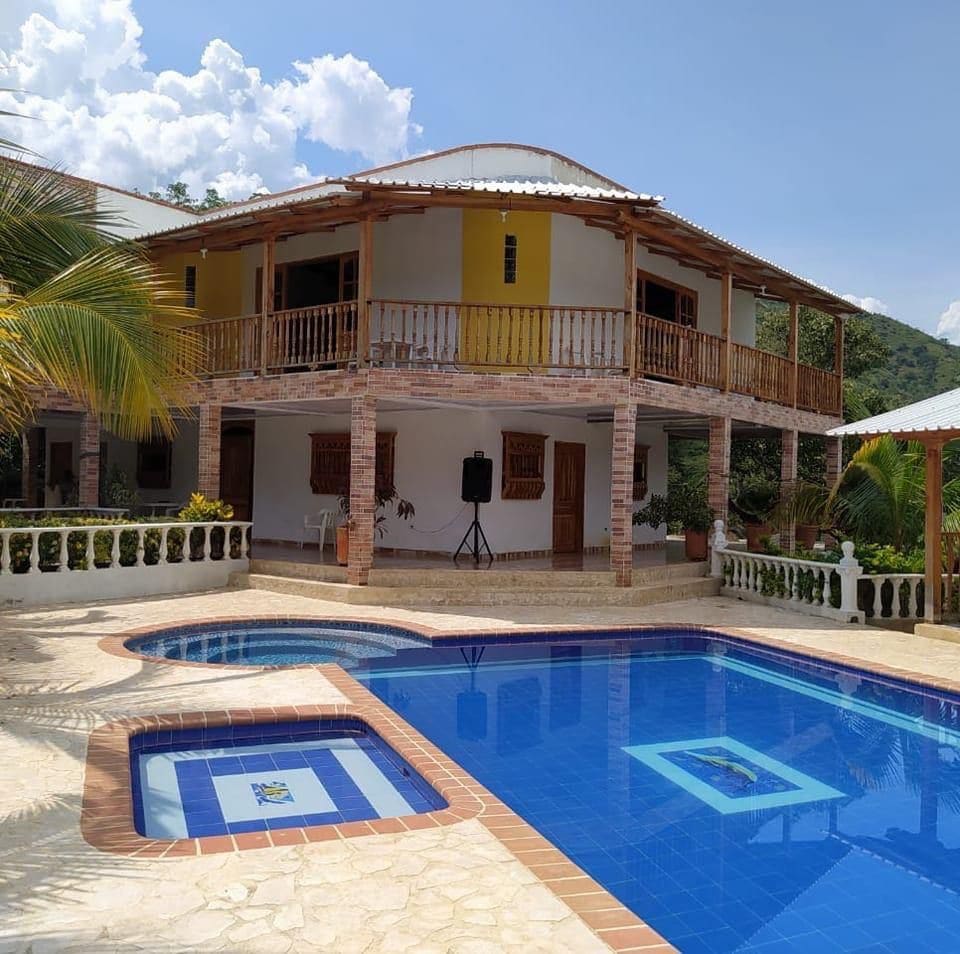 A modern finca is displayed with a large blue swimming pool in the foreground. The two-story structure features a spacious balcony and wide windows, surrounded by palm trees. The pool area includes vibrant tiles, creating a welcoming environment for gatherings.