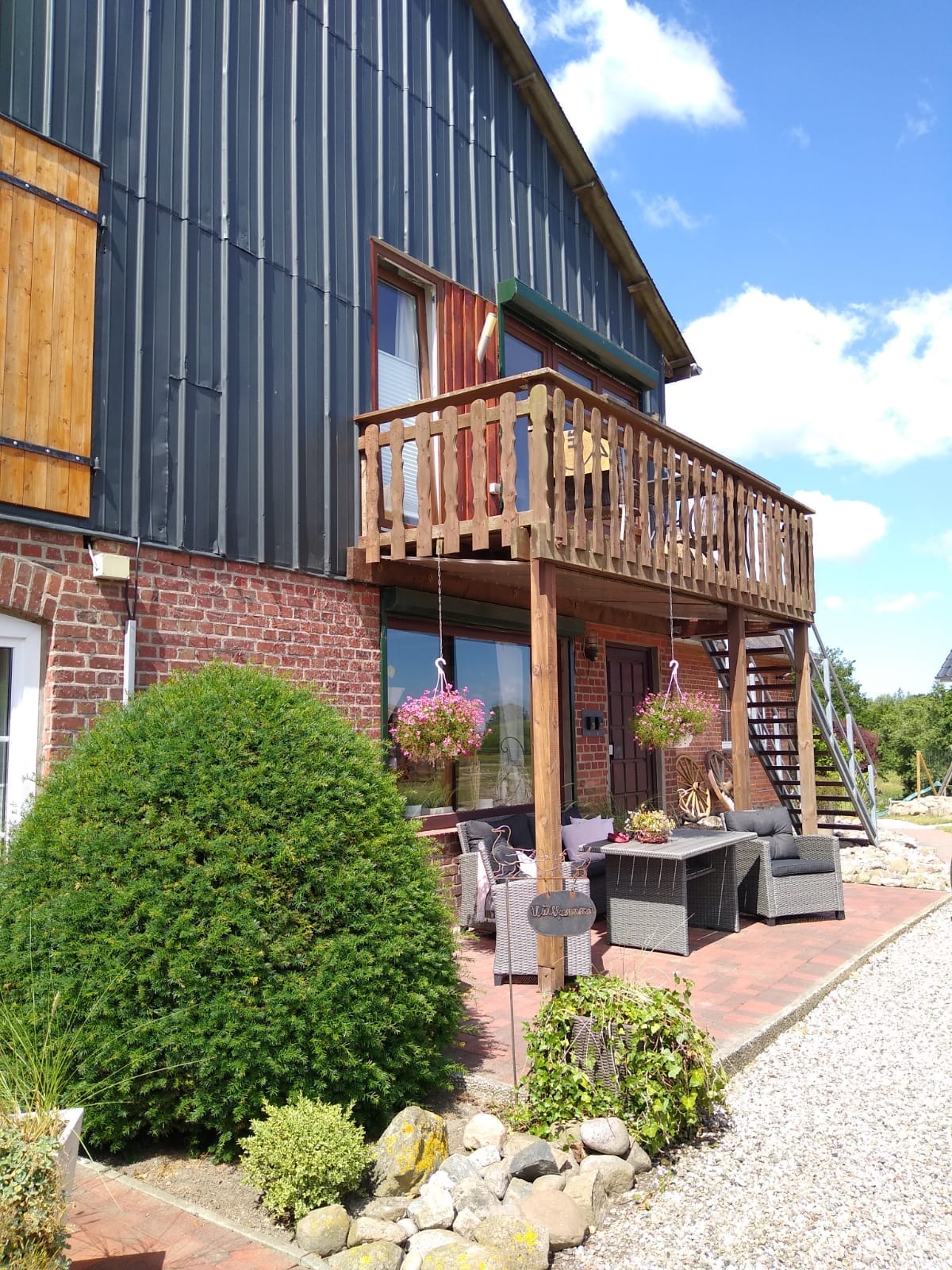 A wooden balcony extends from the upper floor of a rustic building, adorned with hanging flower baskets. Below, a gravel pathway leads to a seating area furnished with comfortable chairs and a small table. Lush greenery and well-maintained landscaping create a welcoming outdoor space.