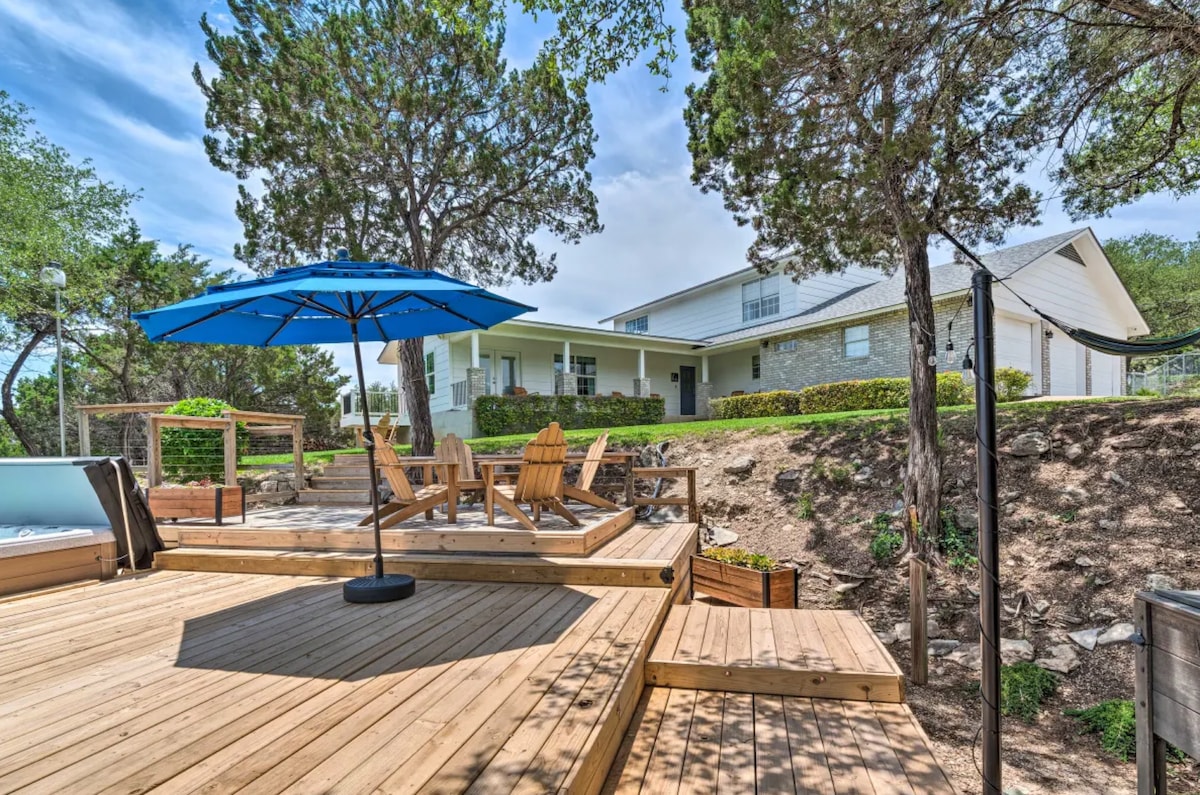 A spacious outdoor deck features wooden flooring with several adirondack chairs and a large umbrella providing shade. In the background, a well-kept villa is visible, surrounded by lush greenery and mature trees.