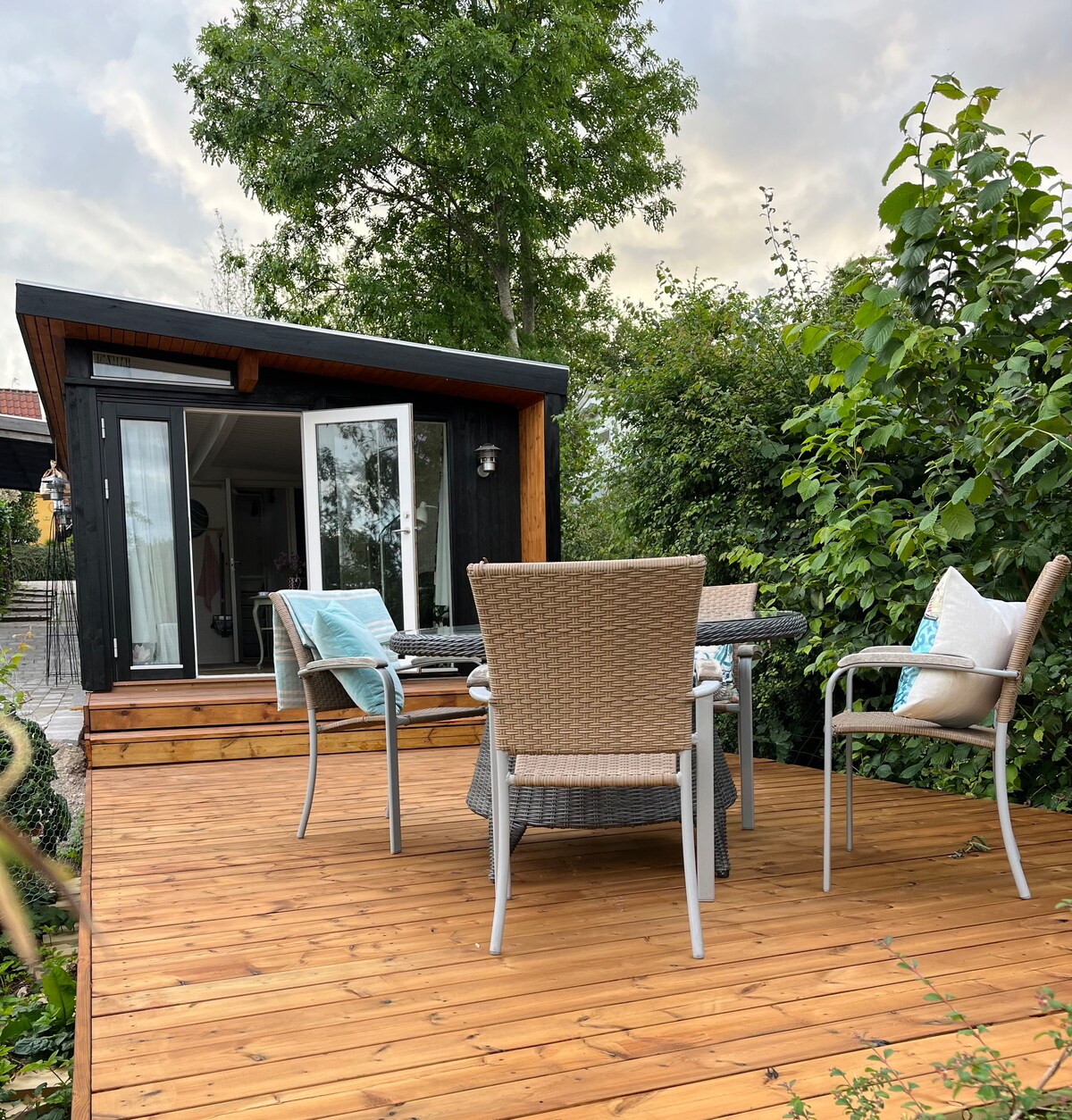 A wooden terrace is surrounded by greenery, featuring a table with four woven chairs. The entrance of the house, framed by large glass doors, provides a glimpse into the inviting interior space.