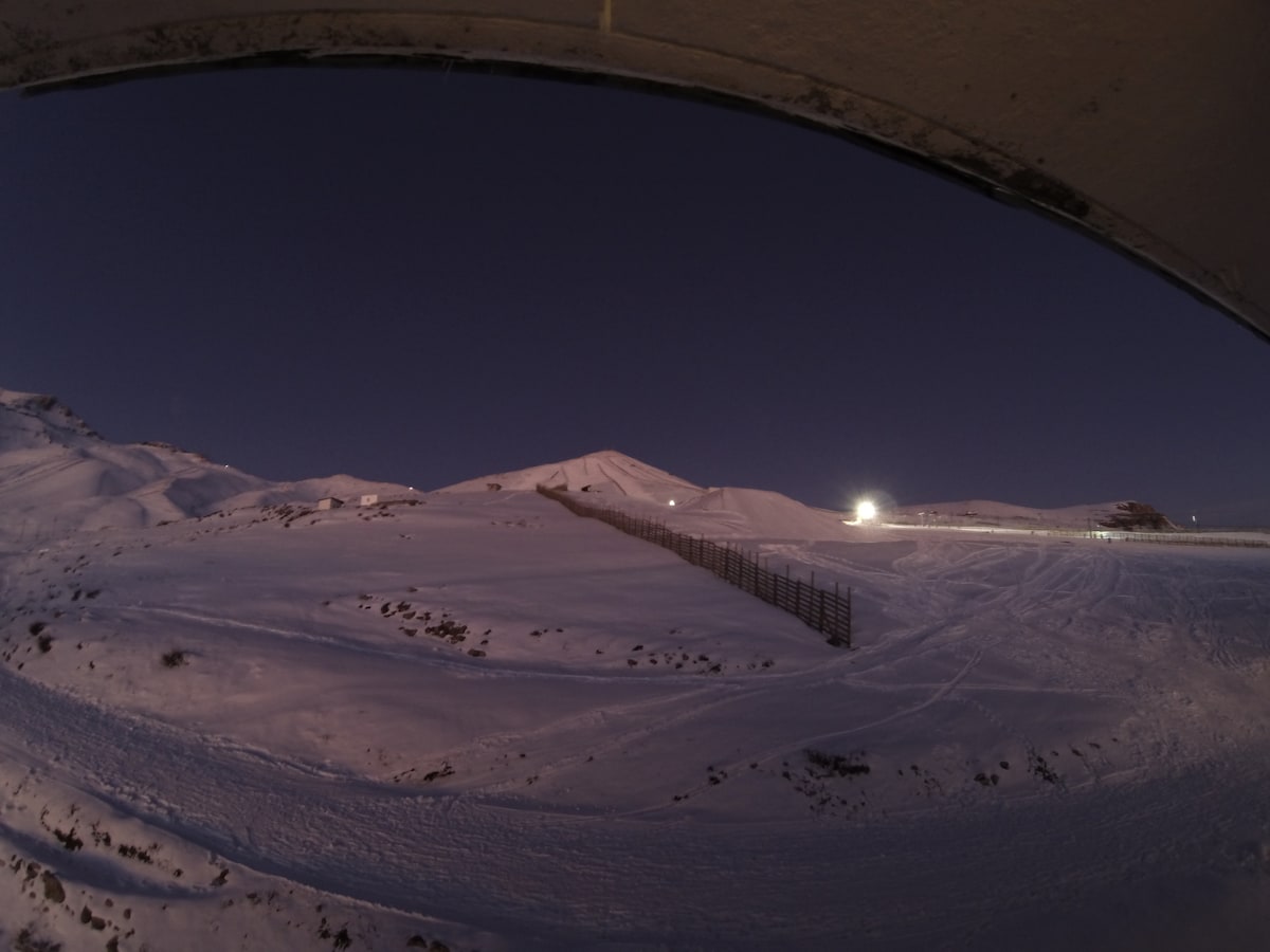 A serene winter landscape is captured under a twilight sky, showcasing a snow-covered slope with a faint outline of ski runs. Soft illumination from ski area lights adds a gentle glow to the scene, while tranquil, untouched snow blankets the terrain.