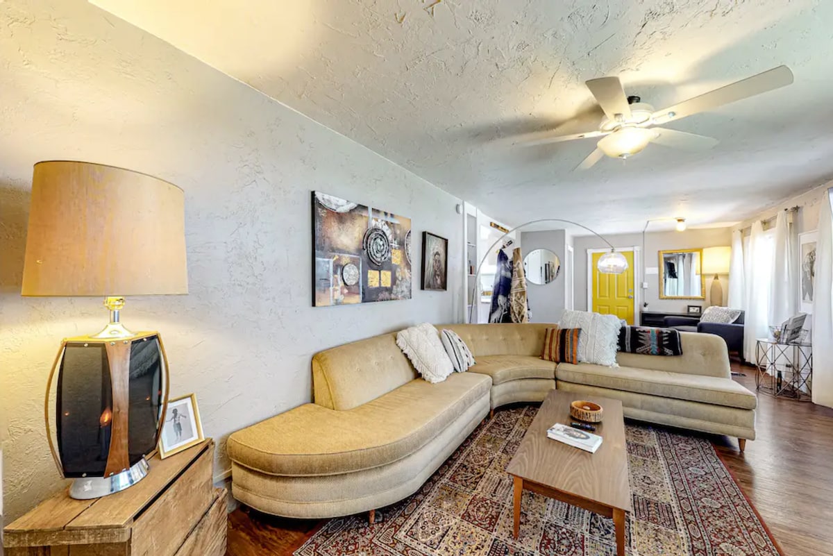 A spacious living area features a large curved sectional sofa adorned with patterned pillows. A wooden coffee table sits atop a decorative area rug, complemented by a lamp with a beige shade. Wall art and a ceiling fan add to the cozy atmosphere, while natural light filters through sheer curtains.